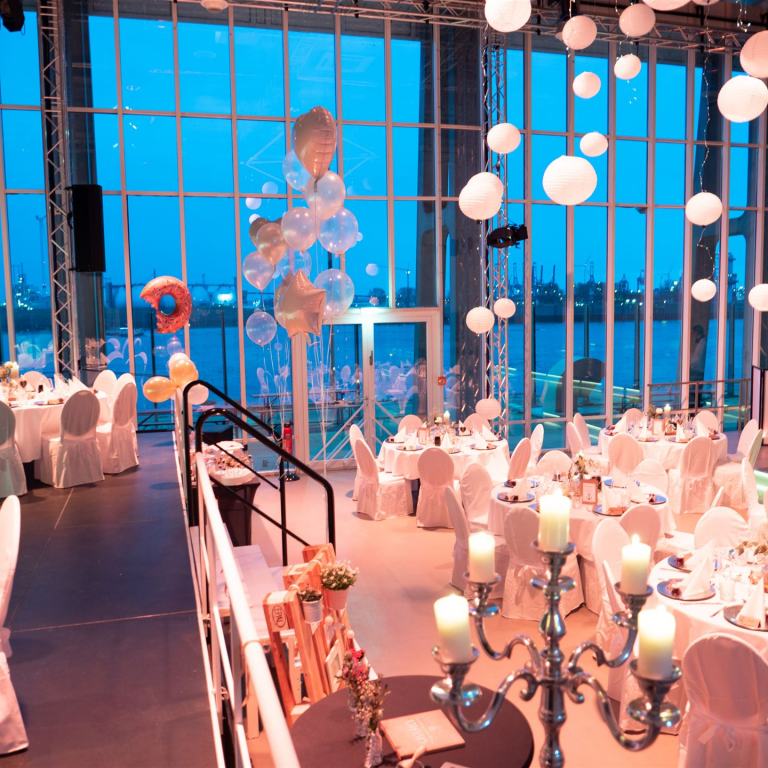 Der Saal ist für eine Hochzeit dekoriert. Im Vordergrund sieht man einen Kronläuchter und im Hintergrund Ballons und weiße Lampignons, die an der Decke hängen. im Saal stehen viele Rundtische.